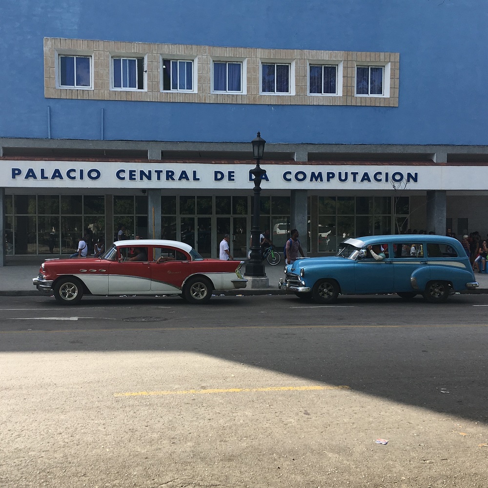 Außenansicht des blauen Gebäudes des Palacio Central de la Computaciòn im 60er Jahre Stil. Vor dem Gebäude parken zwei für das Stadtbild von Havanna charakteristische Oldtimer