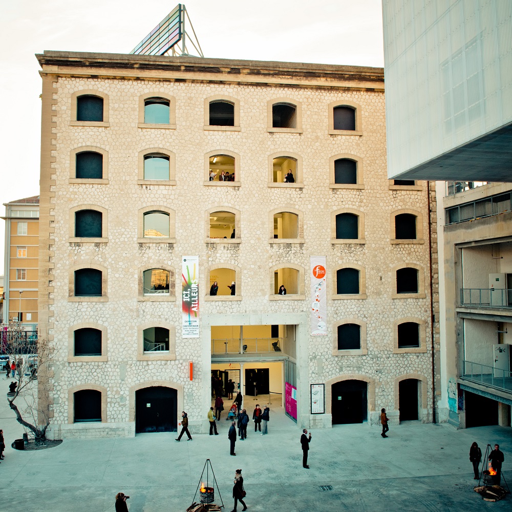 Teilansicht des Kreativstandortes La Friche la Belle de Mai in Marseille. ZU sehen ist ein modern aufgearbeitetes sechsstöckiges historisches Gebäude, das zu Kulturzwecken genutzt wird. Auf dem Platz vor dem Gebäude stehen einige Menschen und unterhalten sich an Feuerschalen.
