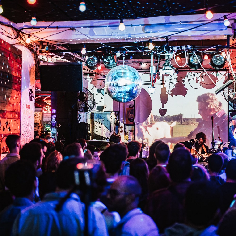 The photo shows a concert scene in the club Kuli Alma in Tel Aviv. From the ceiling hangs a glittering disco ball, in front of a full audience.