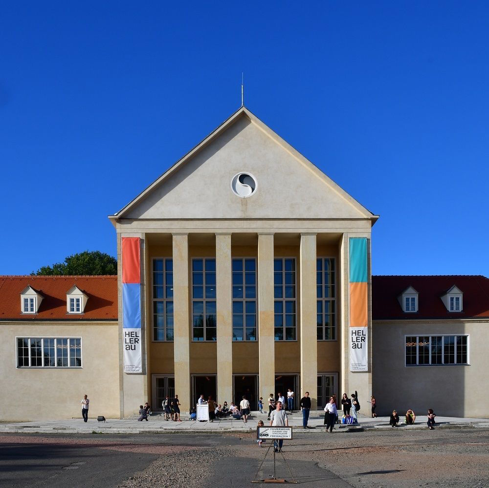 Außenansicht des Festspielhauses Hellerau. Vor dem Gebäude sitzen und stehen mehrere Personen. Der Himmel ist strahlend blau.