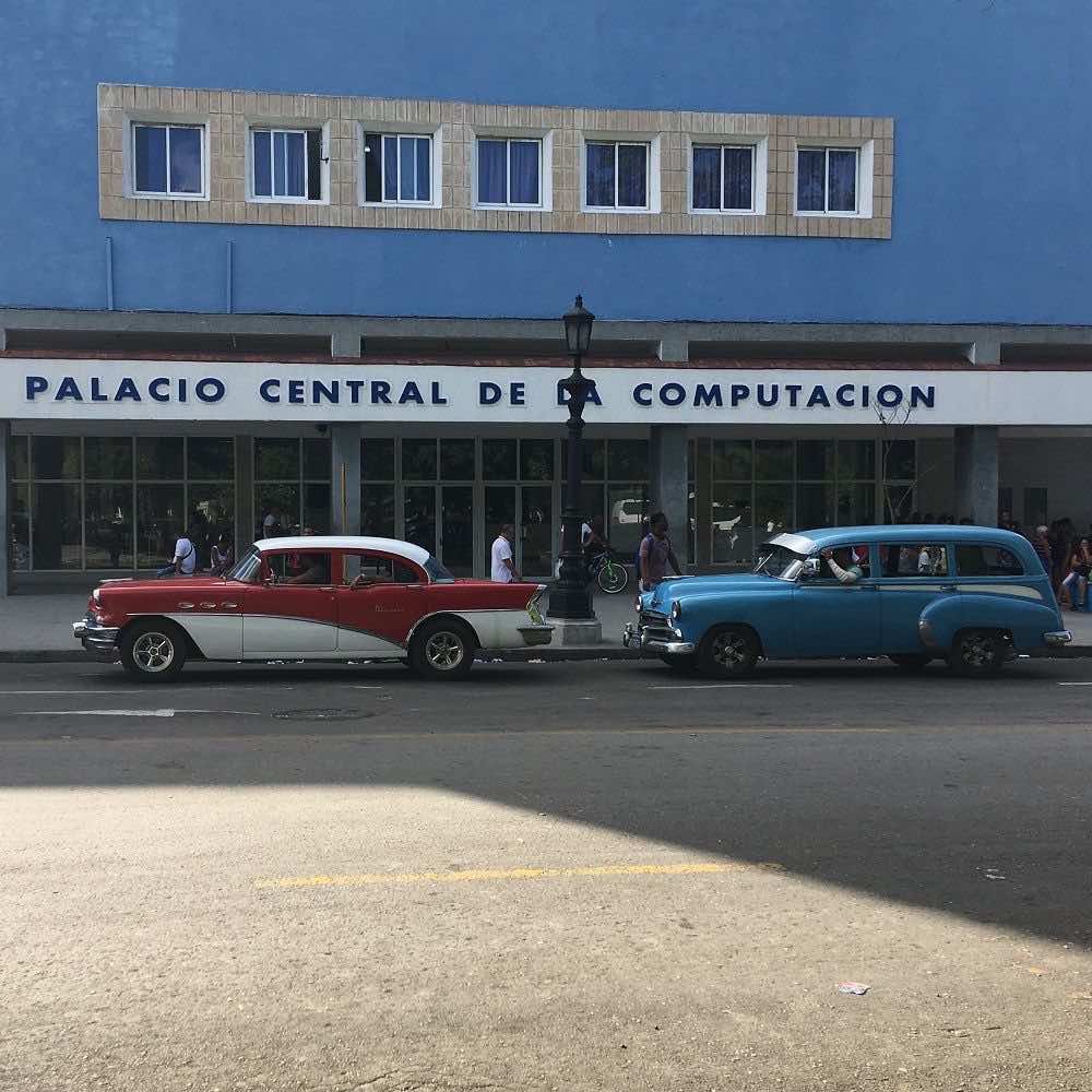 Außenansicht des blauen Gebäudes des Palacio Central de la Computaciòn im 60er Jahre Stil. Vor dem Gebäude parken zwei für das Stadtbild von Havanna charakteristische Oldtimer