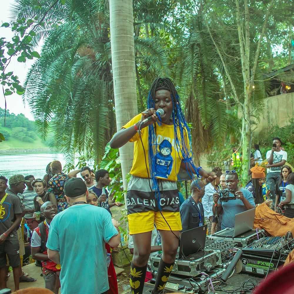 Momentaufnahme im Rahmen des Nyege Nyege Festivals in Jinja, Uganda. Eine Musiker:in mit langen blauen Haaren und einem gelben Outfit performt auf einer Anhöhe. Im Hintergrund sind Tontechnik, ein diverses Publikum, Palmen und ein Flussufer abgebildet.
