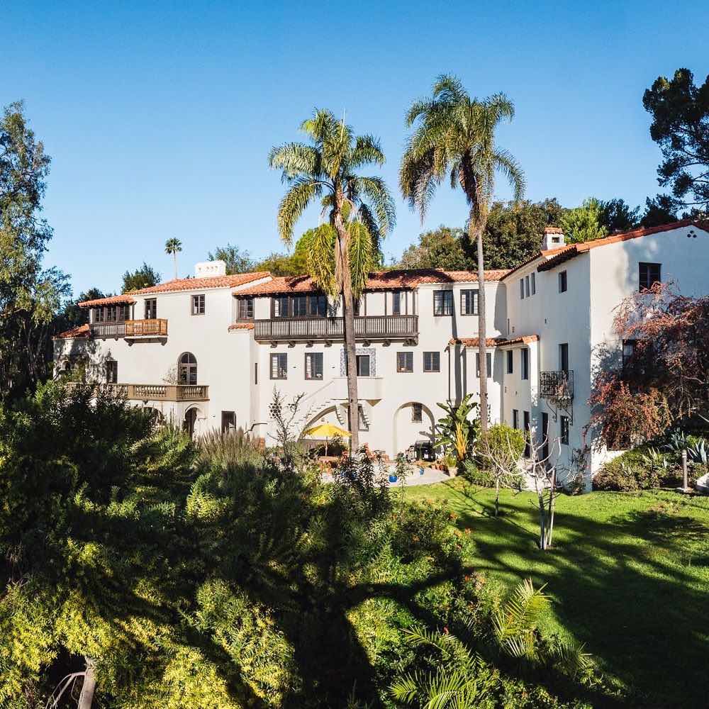 Außenansicht der Villa Aurora in Los Angeles bei strahlend blauem Himmel. Die Schatten der Bäume und Palmen vor dem großzügigen, herrschaftlichen Gebäude werden Schatten auf die vor der Villa liegende Parkanlage.