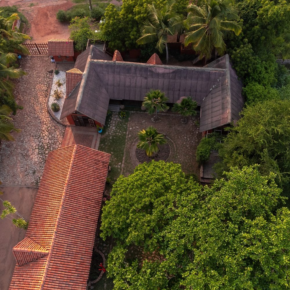 Bird's eye view of the Kokrobitey Institute in Accra. In the photo, there are two buildings that are L-shaped to each other. They are surrounded by lush green trees and palm trees. On the left side is a sandy, rust-colored driveway leading to the site.