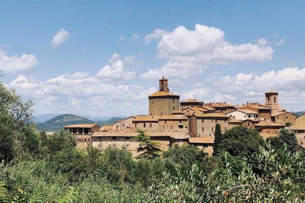 View of the medieval old town of Panicale. The sky is blue, only a few clouds cover it. In the foreground are Mediterranean bushes.
