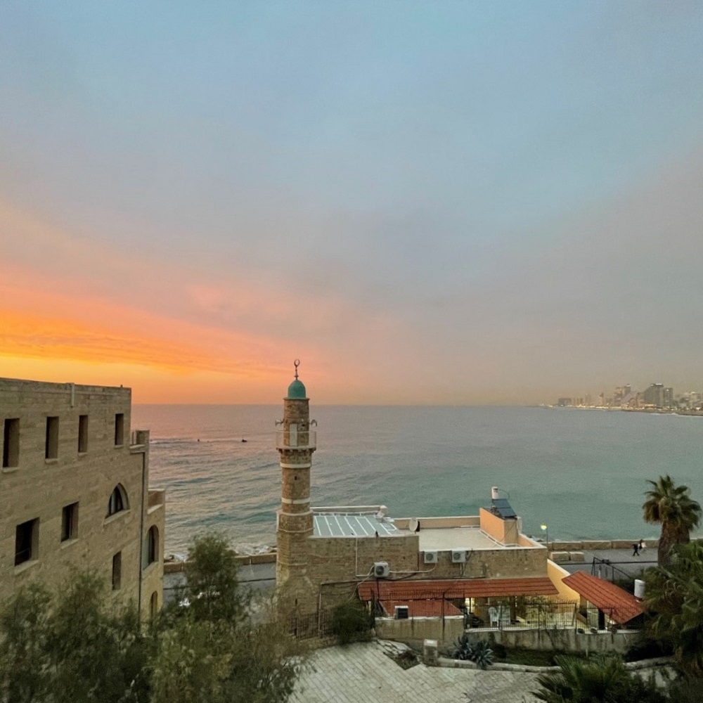 The photo shows a view of the Yafo district of Tel Aviv. On the right side, the skyline of downtown Tel Aviv can be seen in the distance. In the center of the photo is a mosque, which is a few meters away from the sea. The sun is setting over the sea.