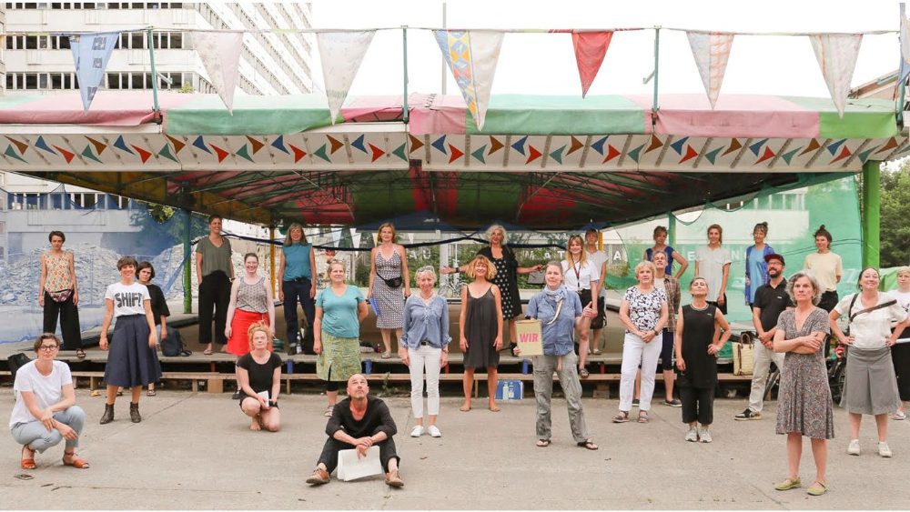 The photo shows about 30 people standing in 3 rows, spaced apart in front of the bumper cars at the Haus der Statistik in Berlin-Mitte. The people, seemingly a choir formation, wear clothes in a wide variety of colors and styles.