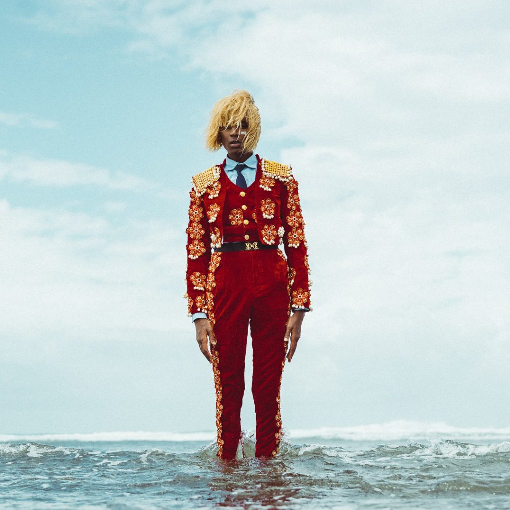 Kabeaushé wearing a red velvet suit with flowers, standing in a light blue ocean