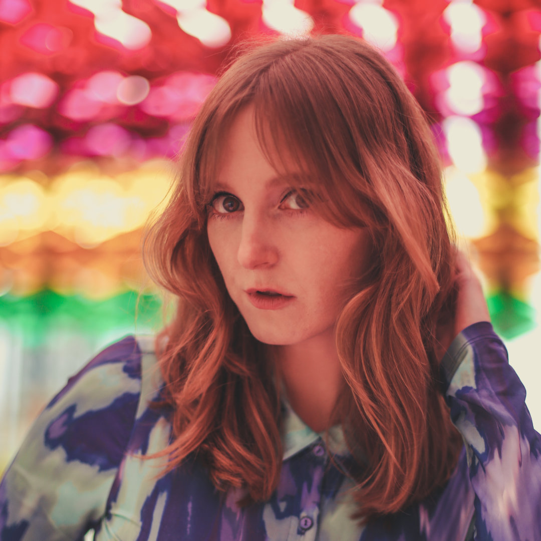 Portrait of Sara. She is wearing a pattern blouse and is looking directly at the camera. Blurred, colorful lights in the background.