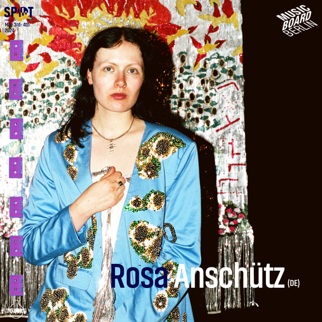 Rosa in a light blue shiny blazer embroidered with sequins, looking directly into the camera. In the background an artfully crafted curtain. The image also depicts her name (Rosa Anschütz) and the SPOT Festival and Musicboard logos.